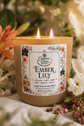 Essence Cove Ember Lily 10 oz luxury olive oil wax candle with double cotton wicks and gold lid lit on a stone surface surrounded by lilies and orchids.